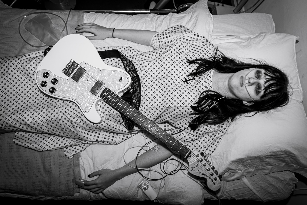 Grace lying down wearing a medical gown in a hospital. Her eyes are closed, and her guitar is resting on top of her.